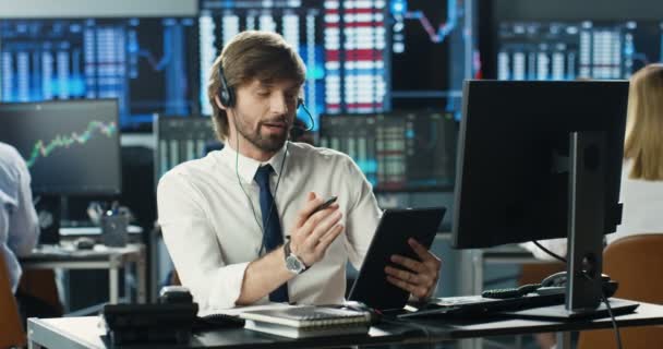 Homem caucasiano corretor no fone de ouvido sentado na frente do monitor do computador falando com o cliente. Batendo no dispositivo tablet. Comerciante masculino falando, comprando, vendendo ações. Centro de atendimento. Apoio ao cliente — Vídeo de Stock