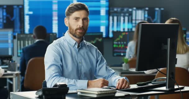 Portrait of young Caucasian handsome man trader working at computer, looking at camera and smiling in trading office of stock market. Happy cheerful male successful broker with smile. Businessman. — Stock Video