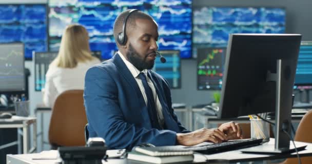 Hombre afroamericano guapo con auriculares. Corredor trabajando en el monitor de la computadora y hablando con el cliente. Hombre comerciante de acciones hablando por teléfono en la oficina de comercio y escribir en el teclado. Departamento de ventas. — Vídeos de Stock