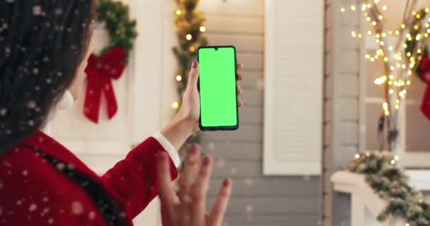 Ferme là. Vue sur l'épaule d'une femme caucasienne tenant un smartphone avec écran vert et agitant la main debout à l'extérieur la veille de Noël. Téléphone portable avec clé chromatique dans les mains de la femme. Concept d'appel vidéo — Video