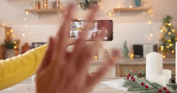Primer plano de la mano femenina caucásica sosteniendo el teléfono inteligente y hablando en videollamada en línea con amigos de razas mixtas y familiares que tienen múltiples chat en gadget en Año Nuevo en casa. Celebrando la Navidad — Vídeos de Stock