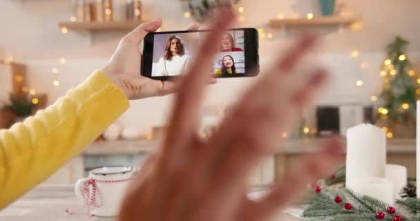 Женщина держит в руках смартфон с онлайн видео-звонком, разговаривая с многонациональными азиатскими и кавказскими друзьями, празднующими Новый год на украшенной кухне. Зимние каникулы. Закрывай. несколько чатов — стоковое видео