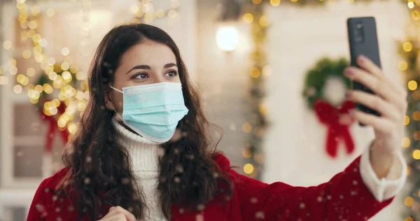 Medikal maskeli neşeli beyaz kadının portresini yakın çekim yapın. Açık havada süslenmiş noel sokağında dururken akıllı telefondan görüntülü konuşma yapıyor. Şehirde kar yağıyor. Mutlu yıllar konsepti