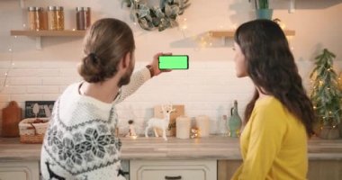 Kafkasyalı genç ve mutlu çiftin arka planında süslü evde oturan ve ellerinde yeşil ekranlı akıllı telefonları tutan bir erkek ile internette konuşan kadın ve erkek. Krom anahtar. Noel konsepti