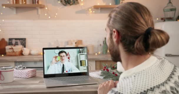 Sobre la vista del hombro del tipo caucásico sentado en casa decorada hablando en video llamada en línea en el ordenador portátil con el médico masculino en cuarentena en la temporada de Navidad. Médico mostrando pastillas en la consulta de vídeo — Vídeos de Stock