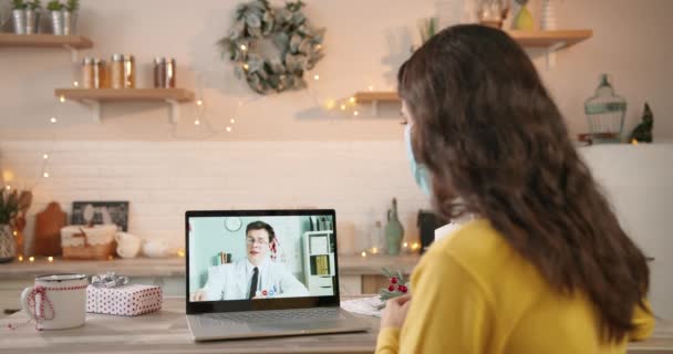 Au-dessus de l'épaule. Gros plan de femme caucasienne dans le masque médical dans la cuisine décorée ayant chat vidéo en ligne sur ordinateur portable avec un médecin masculin en quarantaine pendant la période de Noël. Médecin consulter femme en ligne — Video