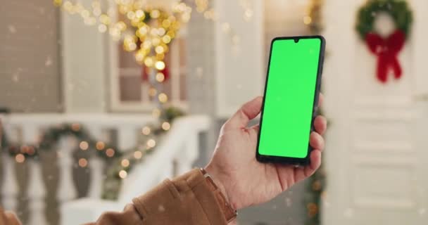 Close up shot of Caucasian male hand holding black smartphone with green screen while standing outdoors on Christmas Eve. Cellphone with chroma key in man hands outdoors in winter. Technology concept — Stock Video