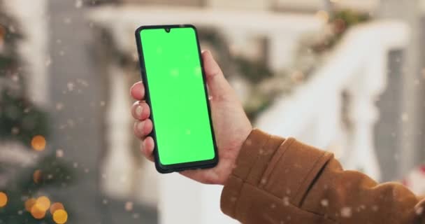 Κοντινό πλάνο του μαύρου smartphone με πράσινη οθόνη στο λευκό ανδρικό χέρι, ενώ στέκεται έξω την παραμονή των Χριστουγέννων. Κινητό τηλέφωνο με chroma κλειδί σε ανθρώπινα δάχτυλα το χειμώνα. Τεχνολογική έννοια — Αρχείο Βίντεο