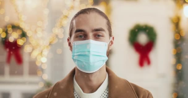 Gros plan portrait de joyeux jeune homme beau en masque médical debout sur la rue décorée christmassy en ville, regardant la caméra et souriant au Nouvel An. Joyeuses fêtes en quarantaine. Concept de Noël — Video