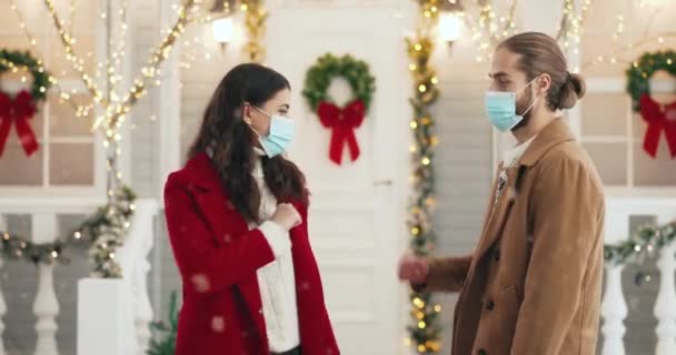 Portrait of happy Caucasian young couple in masks meeting on street in city on New Year while snowing greeting with elbows and speaking. Man and woman friends in festive mood on xmas in quarantine — Stock Video