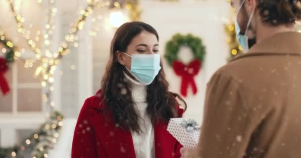 Ritratto ravvicinato di bella donna caucasica felice in maschera medica che riceve il regalo di Natale da uomo amato durante Natale su strada nevosa. Amare marito dando regalo a moglie all'aperto il nuovo anno — Video Stock