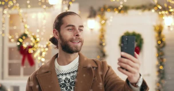 Κοντινό πλάνο πορτρέτο του χαρούμενου Καυκάσου νεαρού όμορφου άνδρα σε εορταστική διάθεση στέκεται σε διακοσμημένο δρόμο με φώτα λάμψης και μιλώντας σε βίντεο chat στο smartphone. Ευτυχισμένο το Νέο Έτος Γιορτές — Αρχείο Βίντεο