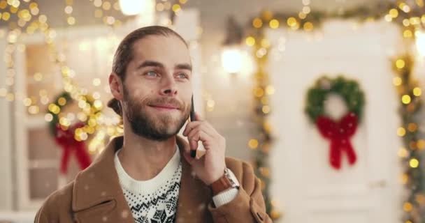 Close up portrait of happy Caucasian young handsome man in festive mood standing in decorated street with twinkle lights and calling on smartphone while snowing. Happy New Year. Xmas concept — Stock Video