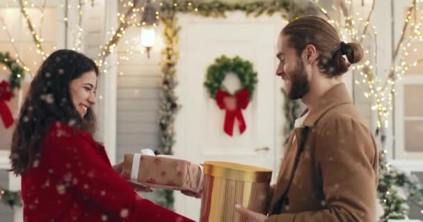 Close up portrait of cheerful Caucasian young married couple exchanging xmas presents while standing on decorated snowy street. Side view. Husband receives and opens gift box with surprised face. — Stock Video