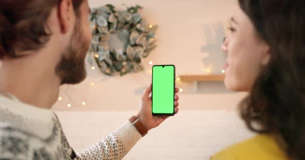 Achteraanzicht van Kaukasisch gelukkig getrouwd jong stel man en vrouw in versierd huis praten op video terwijl de man houdt mobiele telefoon met groen scherm in handen. Chroma sleutel. Kerstconcept — Stockvideo