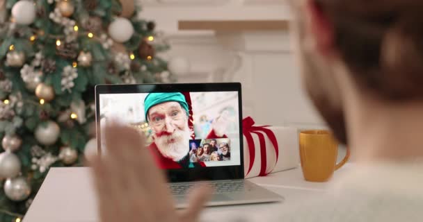 Back view of happy man waving hand and videochatting on laptop with his Caucasian family and sending xmas greetings online. Guy talking on video call with old dad in Santa Claus costume and relatives — Stock Video