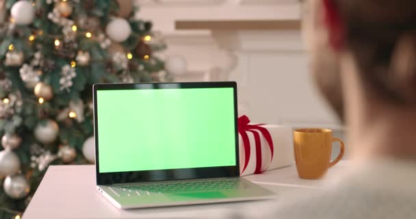 Przez ramię widok młodego człowieka siedzącego w urządzonym pokoju i patrząc na laptopa z zielonym ekranem na Boże Narodzenie. Biały mężczyzna w Wigilię przy użyciu komputera z kluczem chromowym. Zamknij drzwi. Koncepcja techniczna — Wideo stockowe