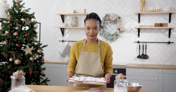 Mulher feliz em avental de pé à mesa em casa cozinha acolhedora e segurando bandeja com biscoitos xmas adicionar biscoitos decorados. Afro-americano belo cozimento feminino na véspera de Natal. Conceito de retrato — Vídeo de Stock