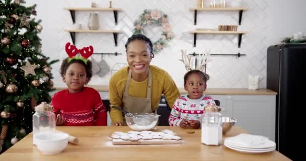 Afro-americano mamma gioiosa con i bambini piccoli abbastanza in piedi a tavola in cucina accogliente a casa, mentre facendo biscotti di Natale e biscotti. Famiglia felice che sorride alla telecamera la vigilia di Natale. Concetto di Capodanno — Video Stock