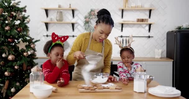 Heureuse famille afro-américaine avec de petits enfants mignons à Noël debout à la table dans la cuisine et décorer les biscuits maison au pain d'épice de Noël Préparatifs du Nouvel An, concept de vacances d'hiver — Video
