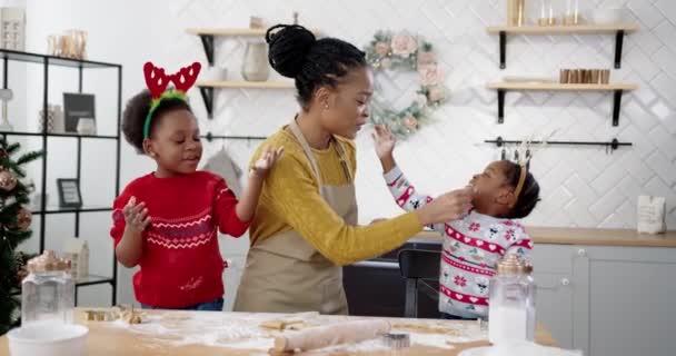 Wesoła Afroamerykanka w fartuchu z małymi dziećmi stojącymi przy stole w domu Boże Narodzenie urządzone kuchnia i zabawy podczas robienia ciasteczek świątecznych. Koncepcja urlopu — Wideo stockowe
