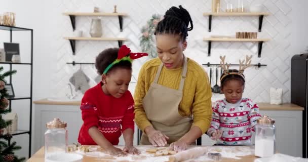 Ritratto di mamma afro-americana sorridente in grembiule con bambini piccoli a tavola in casa cucina decorata di Natale e divertirsi mentre si fanno i biscotti di Natale. Felice concetto olandese — Video Stock
