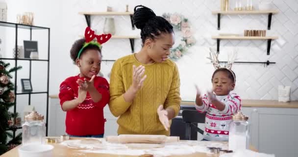 クリスマスの装飾されたキッチンにテーブルに立っている小さなかわいい子供たちと楽しいアフリカ系アメリカ人のお母さんは、自宅でクリスマスクッキーを作りながら楽しんでいます。新年の準備。休日のコンセプト — ストック動画