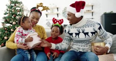 Noel Baba şapkalı mutlu Afrikalı Amerikalı babanın Noel ağacının yanındaki süslü evde otururken ailesine Noel hediyesi verdiği portresi. Yeni yıl kutlaması, Noel hediyeleri. Tatil kavramı