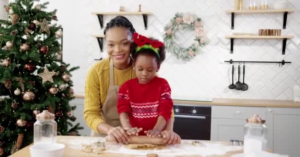 Πορτρέτο της ευτυχισμένης αφρικανικής αμερικανικής μαμάς με μικρό χαριτωμένο παιδί σε χριστουγεννιάτικο πουλόβερ στέκεται στο τραπέζι στο σπίτι διακοσμημένα κουζίνα και χαμογελώντας στην κάμερα, ενώ κάνοντας ζύμη για τα μπισκότα gingerbread Χριστούγεννα — Αρχείο Βίντεο