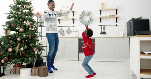 Il papà afroamericano e il bambino ascoltano musica natalizia e ballano nella moderna cucina di casa decorata con l'albero di Capodanno la vigilia di Natale. — Video Stock