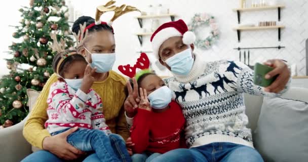 Portrait of happy African American family in masks taking pictures on smartphone in cozy christmassy decorated home. Dad in santa hat taking selfie photo on cellphone with kids and wife — Stock Video