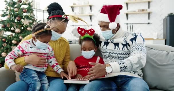 Afroamerikanische Familie in Masken mit Kindern, die zu Hause am Weihnachtsbaum sitzen und Bücher lesen. Neujahrsfeier in Quarantäne. Eltern und Kinder verbringen Zeit miteinander — Stockvideo