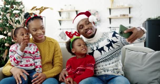 Portrét šťastné afroamerické rodiny fotografující na smartphonu a usmívající se v útulné vánoční zdobené domů. Táta v Santa klobouku fotit selfie na mobilu s dětmi a manželkou — Stock video