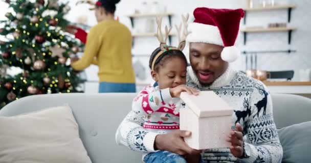 Portrait of curious African American joyful kid opens gift box while sitting with happy dad in santa hat together at decorated home. Mom decorating Christmas tree on background. Xmas presents concept — Stock Video