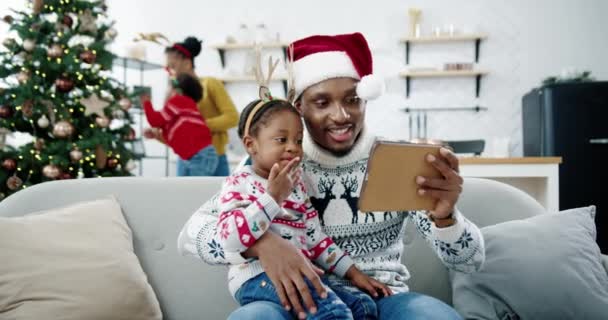 Primo piano di felice bambino afroamericano seduto con papà in cappello di Babbo Natale nella stanza decorata e guardare cartoni animati su tablet. Mamma e bambino che decorano l'albero di Natale sullo sfondo. Concetto di Natale — Video Stock