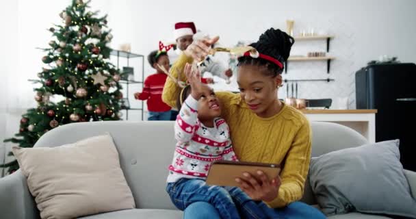 Nahaufnahme einer glücklichen afroamerikanischen Familie zu Hause. Kleine süße Kind sitzt mit Mama in dekorierten Raum und mit Tablet. Papa und Kind schmücken den Weihnachtsbaum im Hintergrund. Winterferienkonzept — Stockvideo