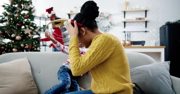 Zbliżenie szczęśliwy Afroamerykanin małe dziecko siedzi z mamą na kanapie w domu Bożego Narodzenia i zabawy. Tata i dzieciak dekorowali choinkę na tle. Duch Nowego Roku Grudniowa koncepcja wakacji — Wideo stockowe