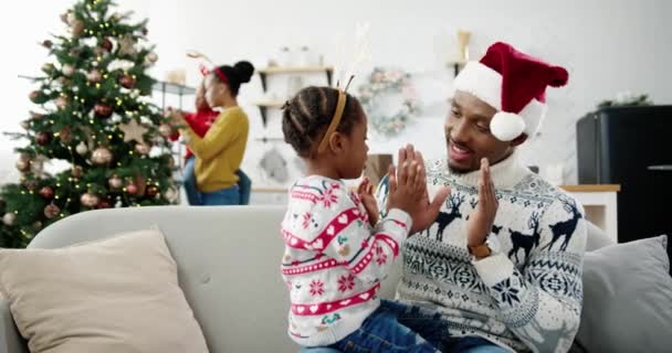 Close up retrato de pai ensinando criança pequena uma canção e brincando enquanto sentado no sofá na casa de Natal. Afro-americano mãe e criança decoração árvore de Ano Novo no fundo. Conceito de Natal — Vídeo de Stock