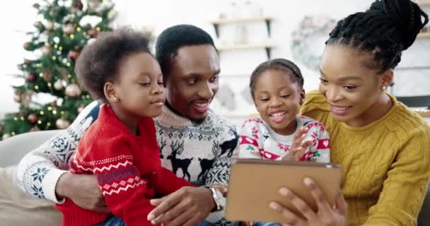 Primo piano di felice famiglia afroamericana a casa seduta in camera decorata vicino all'albero di Natale e utilizzando tablet Piccoli bambini carini seduti con mamma e papà e toccando tablet scegliere regali online — Video Stock