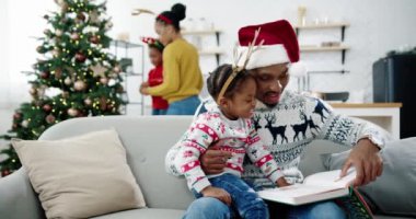 Küçük mutlu çocuğa kitap okuyan Afrikalı Amerikalı neşeli babanın portresi rahat modern evdeki kanepede otururken. Arka planda çocuk süsleme ağacı olan güzel bir anne. Aile kavramı