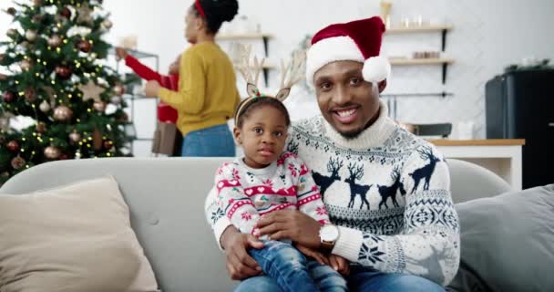 Portrét Afroameričana veselého otce s malým roztomilým dítětem a s úsměvem na kameru, zatímco sedí v útulném moderním domě. Máma a dítě zdobí novoroční stromek na pozadí. Vánoční koncept — Stock video