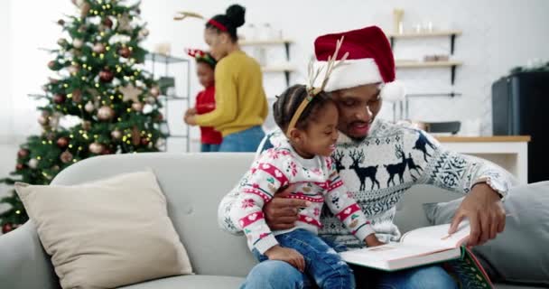 Küçük mutlu çocuğa kitap okuyan Afrikalı Amerikalı neşeli babanın portresi rahat modern evdeki kanepede otururken. Arka planda çocuk süsleme ağacı olan güzel bir anne. Aile kavramı — Stok video