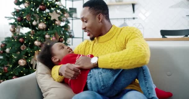 Retrato de feliz padre afroamericano hablando y jugando con un niño pequeño mientras está sentado en el sofá en la habitación decorada navideña Hermoso padre amoroso y el niño pasar tiempo juntos Concepto de vacaciones — Vídeos de Stock