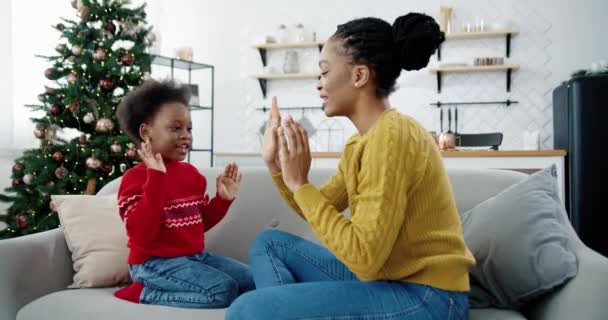 Portret szczęśliwej pięknej Afroamerykanki bawiącej się z małym dzieckiem, siedzącej na kanapie w urządzonym pokoju Bożonarodzeniowym Widok z boku kochającej mamy i dziecka spędzającego razem czas Koncepcja świąt — Wideo stockowe