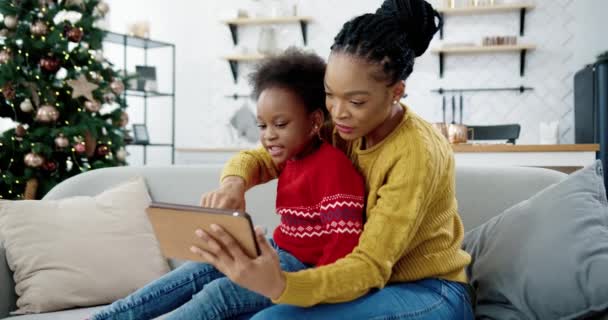 Afroamerikansk glad mamma och barn sitter på soffan i dekorerat modernt rum nära julgran och tittar på något på surfplattan. Kvinnan knackar på enheten och visar den för barnet. December helgdagar — Stockvideo