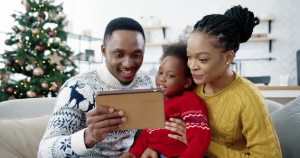 Portret van Afro-Amerikaanse vrolijke vader en moeder met kind zittend op de bank in gedecoreerde moderne kamer in de buurt van kerstboom en kijken naar iets op tablet terwijl kind tikken op apparaat. Gezinsvakantie — Stockvideo