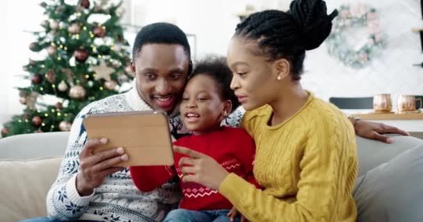 Porträtt av glad afroamerikansk liten unge sitter på soffan i dekorerat rum nära nyårsträd med mamma och pappa och knacka på surfplatta medan du spelar. Familjesemester. God jul konceptet — Stockvideo