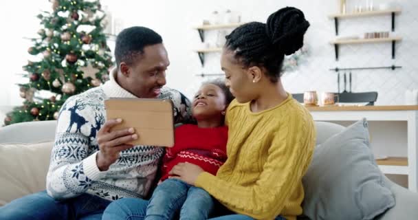 Porträtt av glad afroamerikansk liten unge sitter på soffan i inredda rum nära nyårsträd med mamma och pappa och talar på nätet videosamtal på surfplatta. Familjesemester God Jul koncept — Stockvideo