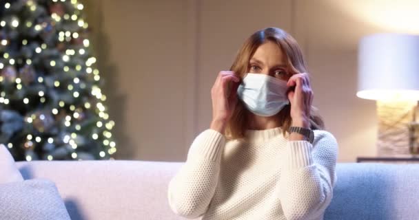 Ritratto ravvicinato di felice donna caucasica che rimanda la maschera medica seduta sul divano in serata a casa vicino all'albero di Natale decorato con luci scintillanti e sorridente alla macchina fotografica. Capodanno — Video Stock
