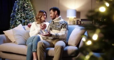 Neşeli Kafkasyalı çift Noel arifesinde parıldayan Xmas ağacıyla rahat bir odada oturuyor. Mutlu koca, evde tatilleri birlikte kutlayan sıcak çay ya da çikolatayla karısıyla konuşuyor.